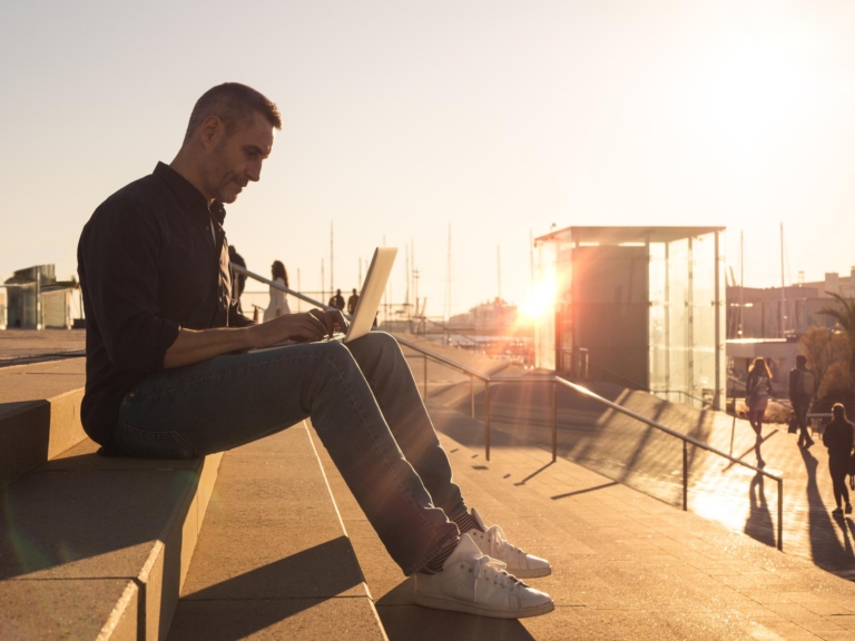 Man sitting on steps outside by water with boats in the background while using a laptop