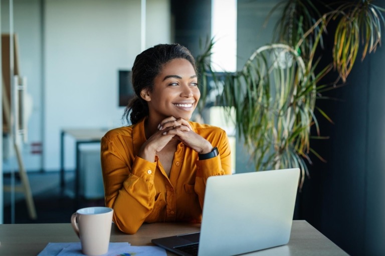 Portrait of businesswoman sitting at desk in office and thinking while working on laptop, looking away with pleased facial expression